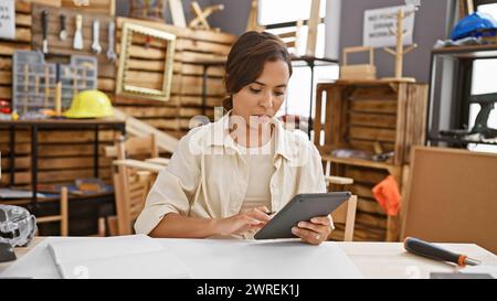 Beautiful young hispanic woman carpenter expertly managing her woodworking business using a touchpad at her indie workshop. Stock Photo