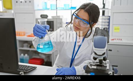 Hispanic woman scientist analyzing blue chemical in laboratory wearing lab coat and safety glasses Stock Photo