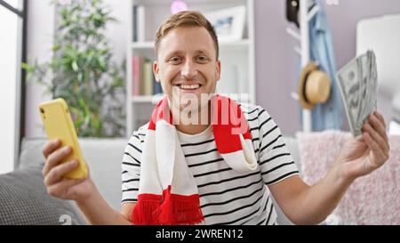 Excited young caucasian man watching his soccer team's game on smartphone at home, holding cash ready for the betting thrill! Stock Photo