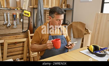 Mature man holding a red mug and smartphone in a woodworking workshop with tools and safety gear. Stock Photo