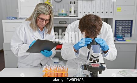 Man and woman, two concentrated scientists together in a lab, sitting, taking notes during microscope analysis Stock Photo