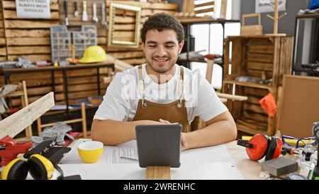 Bearded craftsman in apron using tablet in a carpentry workshop surrounded by tools Stock Photo