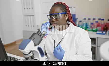 African american woman scientist with braids using microscope and phone in a laboratory setting. Stock Photo