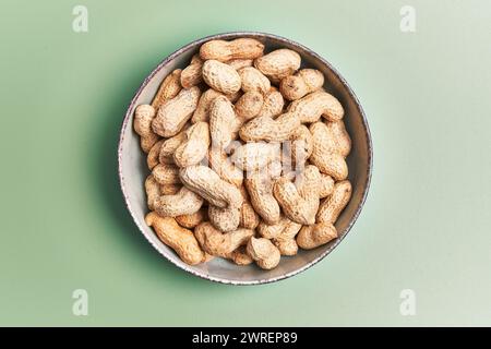 Overhead view of unshelled peanuts in a ceramic bowl on a green background, implying healthy snacking. Stock Photo