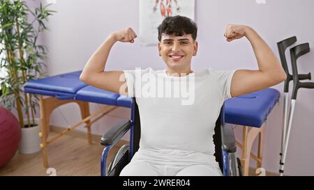 A smiling young man in a wheelchair flexing muscles confidently in a rehab clinic's bright room. Stock Photo