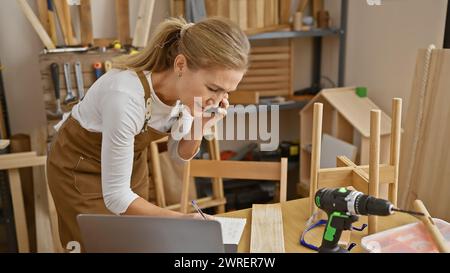 Blonde woman multitasking in a carpentry workshop while talking on a phone and writing notes. Stock Photo