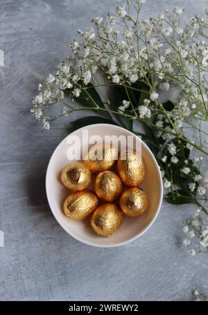 Chocolate Easter Egg Wrapped in Gold Foil. White spring flowers and Ester sweets top view photo. Bright spring still life. Stock Photo