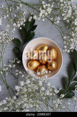 Chocolate Easter Egg Wrapped in Gold Foil. White spring flowers and Ester sweets top view photo. Bright spring still life. Stock Photo