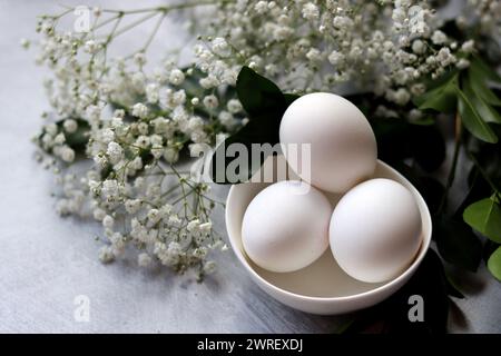 Simple composition still life with eggs and white flowers. Easter celebration concept. Eggs in a white ceramic bowl top view photo. Stock Photo