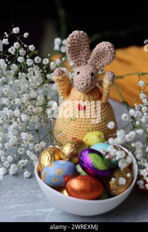 Crochet bunny and bowl of Easter sweets on a table. Colorful still life with chocolate eggs, flowers and toy. Easter celebration concept. Stock Photo