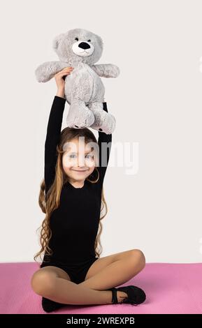 Vertical shot of a little gymnast girl on a yoga mat wearing a black suit, holding her hands up with a teddy bear. High quality photo Stock Photo