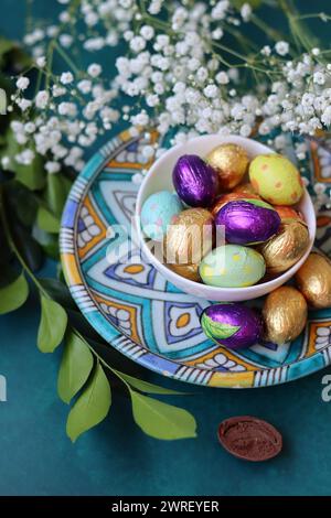 Chocolate Easter Egg Wrapped in Gold Foil. White spring flowers and Ester sweets top view photo. Bright spring still life. Stock Photo