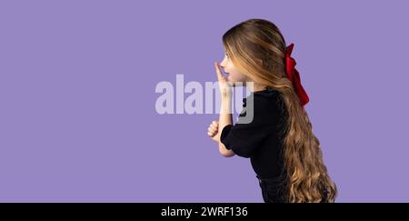 Horizontal shot of cute charming little girl with blond hair wearing black t-shirt posing on purple wall in a studio, looking away with amazed express Stock Photo