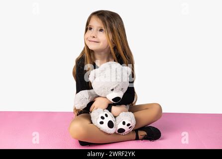 Smiling little girl dressed in black gym clothes, holding a gray teddy bear in her arms, sitting on a pink yoga mat, isolated on a white studio backgr Stock Photo