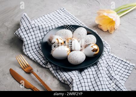 Easter eggs on black plate, checkered napkin and yellow tulip on concrete gray table. Top view. Stock Photo