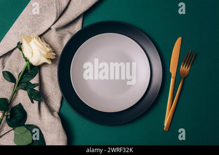 Table place setting with white rose flower, plates and golden cutlery on dark green background. Top view, flat lay. Stock Photo