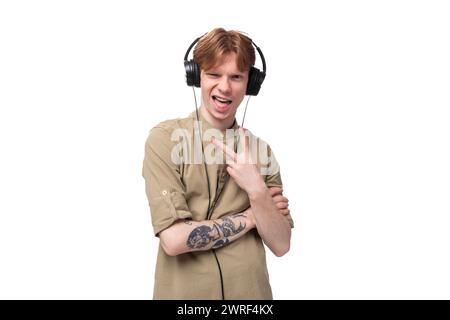 young red-haired man with glasses dressed in a brown shirt grimaces and listens to music on headphones Stock Photo