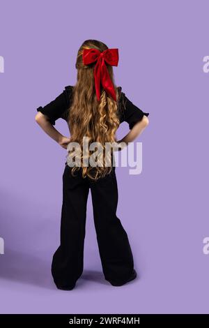 Full back shot of a little girl with long, wavy blonde hair wearing black clothes, hands on hips, isolated on purple background. High quality photo Stock Photo