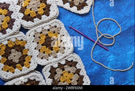 Crocheted granny squares made of natural brown, mustard and beige wool. Soft and fluffy crochet ornament on blue textured background. Stock Photo