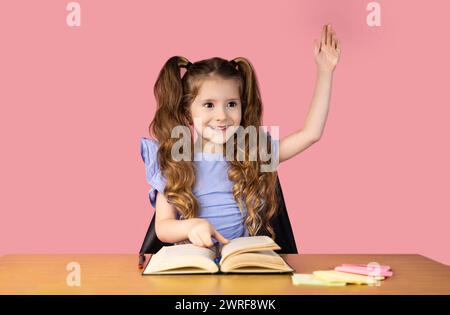 A very beautiful little girl on a pink background, is very active in the lessons and just sits with her hand up to answer the questions given by the t Stock Photo