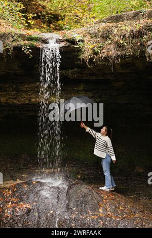young girl standing by the waterfall flowing streams in the morning from a cave low angle holding a black umbrella under the flowing water of the big Stock Photo