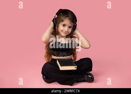 The beautiful little girl with blond hair isolated on a pink background holds a book in her arms and headphones listening to music, concept of relaxat Stock Photo