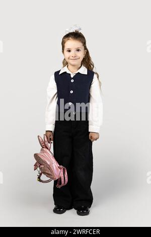 A little girl in a school uniform with an electric blue blazer and collar is holding a pink backpack. She smiles brightly, her thumb hooked through th Stock Photo
