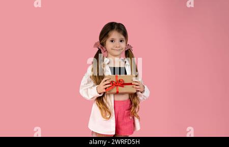 A beautiful little girl on a pink background is holding a gift she received on Valentine's Day. She is very beautiful and smiling, she enjoys this sma Stock Photo