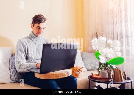 Young man working online from home with pet using laptop. Freelancer typing on computer sitting on couch by ginger cat. Remote job Stock Photo
