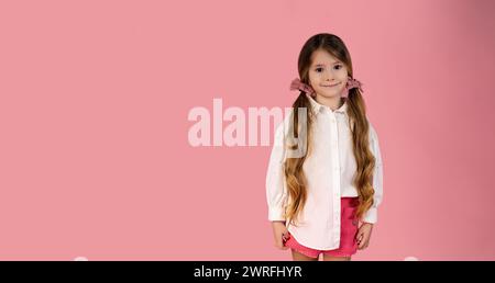 Happy child girl in casual white shirt isolated on pastel pink background with space for text, having pigtails, smiling cheerfully and looking at came Stock Photo