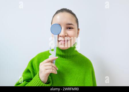 A happy woman in a green sweater is smiling while holding a blue object in front of her eye. Her thumb is resting on the sleeve of her tshirt Stock Photo
