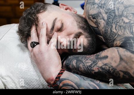 Horizontal photo a man covered in tattoos takes a moment to rest, his hand gently cradling his head, embodying the patience and endurance of getting i Stock Photo