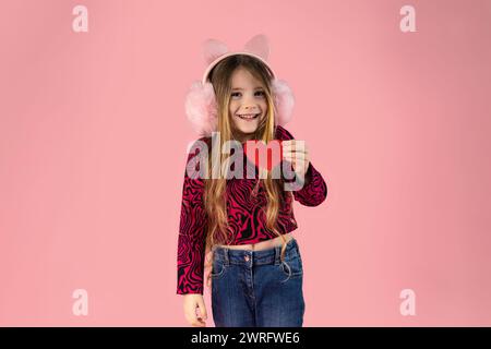 Little girl with long blond hair wearing pink headphones on her ears, dressed in jeans and a pink printed sweater holds a small paper heart and smiles Stock Photo