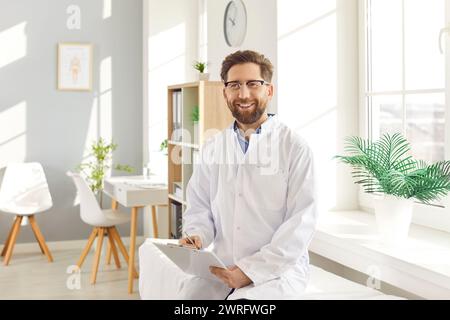 Portrait of a young happy male doctor in white medical uniform sitting on couch in his office. Stock Photo