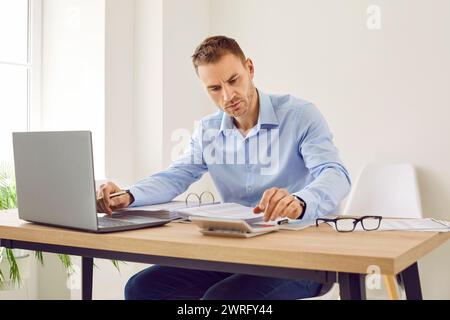 Young focused serious business man working on laptop at office making calculations. Stock Photo
