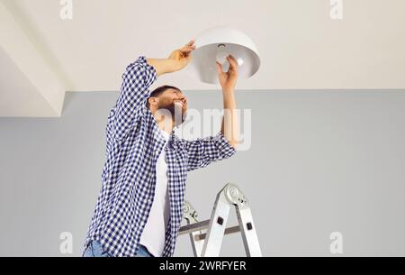 Young bearded man in casual clothes is standing on construction ladder unscrewing burnt-out energy-saving LED light bulb, holding chandelier. Care of Stock Photo