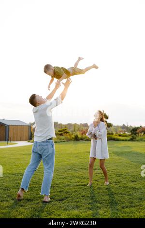 A man is lifting a young boy into the air with his arms extended upwards. The boy is smiling and appears joyful while being held above the ground. Stock Photo