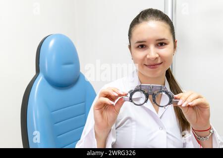 The ophthalmologist is holding a pair of glasses in her hand, with a smile on her face. She is wearing vision care outerwear and gesturing with her ar Stock Photo