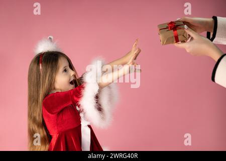 Amazed little girl dressed in red Christmas dress with blond hair receives Christmas present from her mother, isolated on pink background. High qualit Stock Photo