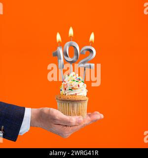 The hand that delivers cupcake with the number 102 candle - Birthday on orange background Stock Photo