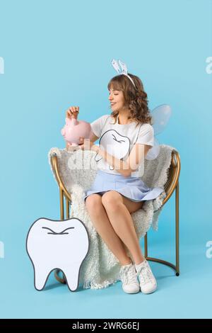 Tooth Fairy putting coin into piggy bank in armchair on blue background Stock Photo