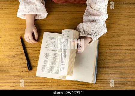 A person is sitting on a wooden floor reading a book placed on a table. Their fingers trace the wooden table, while their other leg rests comfortably Stock Photo