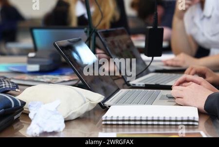 Busy school students working at a table on laptop computers with typical lesson accessories such as chargers, books, pens, rubbish and pencil cases. Stock Photo