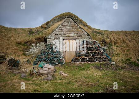 Stone cottage / shed at Sinclairs Bay harbour entrance with lobster and crab pots stacked outside on a sunny day in rural Scotland, ready for fishing Stock Photo