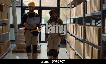 Employee and african american coworker walking through warehouse aisles checking labels on cardboard boxes sitting on shelves, inputing updated products info on online shopping website Stock Photo