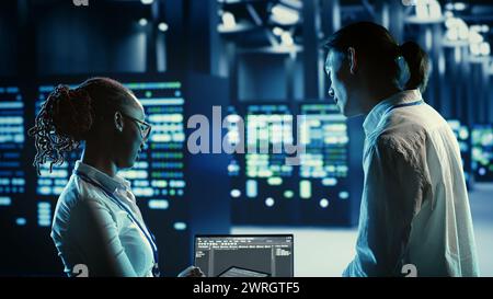 Technicians programming in server room housing advanced infrastructure storing massive datasets. Workers running code on laptop to upgrade data center racks doing complex computational operations Stock Photo
