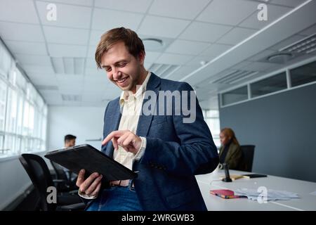 Happy young businessman in businesswear using digital tablet next to desk with coworkers in office Stock Photo
