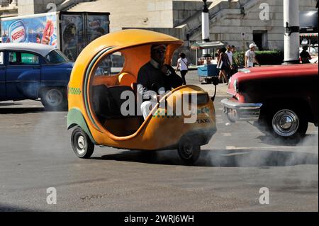 Yellow Coco taxi driving on the street and emitting smoke, vintage car in the background, Havana, Cuba, Central America Stock Photo