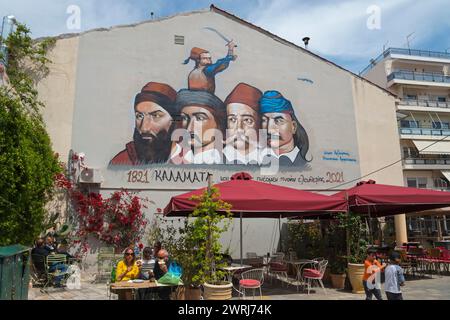 Large mural of historical figures above an urban cafe with passers-by, graffiti with 5 heroes of the Greek Revolution of 1821 on 23 March Square Stock Photo