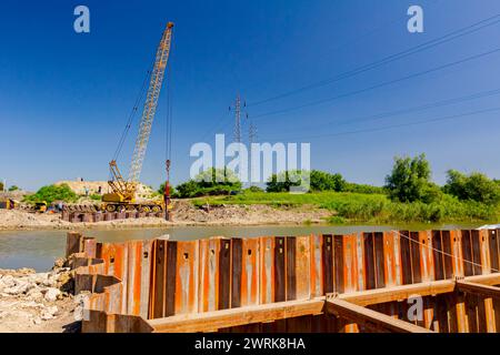 Installed rusty metal piles along river bank built in for bridge foundation at the construction site. Across the water is yellow obsolete mobile crane Stock Photo